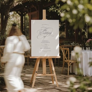 May include: A white rectangular wedding welcome sign with black serif and script typography displayed on a light-colored wooden tripod easel. The text reads: Welcome to The Wedding of Sophia Anderson and Jason Henderson. We're so glad you're here.