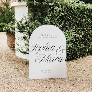 Pu&ograve; includere: Un cartello di benvenuto per matrimoni bianco, ad arco, con un'elegante scritta nera che recita "Benvenuti al matrimonio di Sophia & Marcus". Il cartello include anche il luogo e la data. Il cartello &egrave; all'aperto, circondato dal verde.