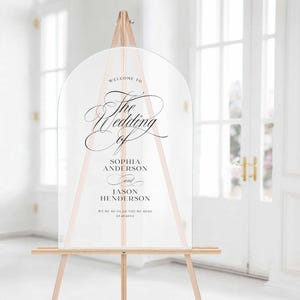 May include: A clear, arched acrylic wedding sign featuring black printed text that reads: Welcome to The Wedding of Sophia Anderson and Jason Henderson. We're so glad you're here 09.25.20XX. The sign is displayed on a light-colored wooden tripod easel.