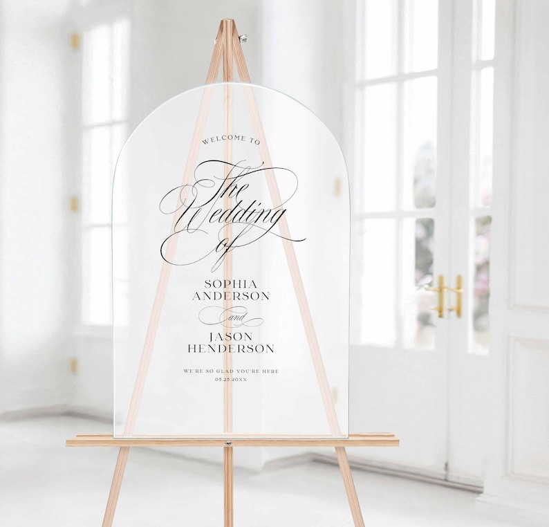 May include: A clear, arched acrylic wedding sign featuring black printed text that reads: Welcome to The Wedding of Sophia Anderson and Jason Henderson. We're so glad you're here 09.25.20XX. The sign is displayed on a light-colored wooden tripod easel.
