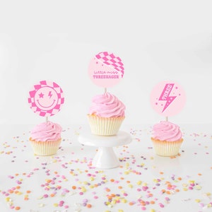 May include: Three cupcakes with pink frosting and decorative toppers. The toppers feature a smiley face, a checkered flag, and the word "THREE" with a lightning bolt. The text "little miss THREENAGER" is also visible. Colorful confetti is scattered around the cupcakes.