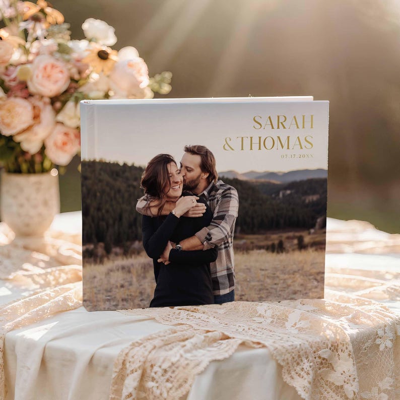 Peut inclure: Un album photo blanc avec une photo d'un couple s'embrassant en ext&eacute;rieur. La couverture porte l'inscription dor&eacute;e "SARAH & THOMAS" et la date "07.17.2023". L'album est pos&eacute; sur une table avec une nappe en dentelle.