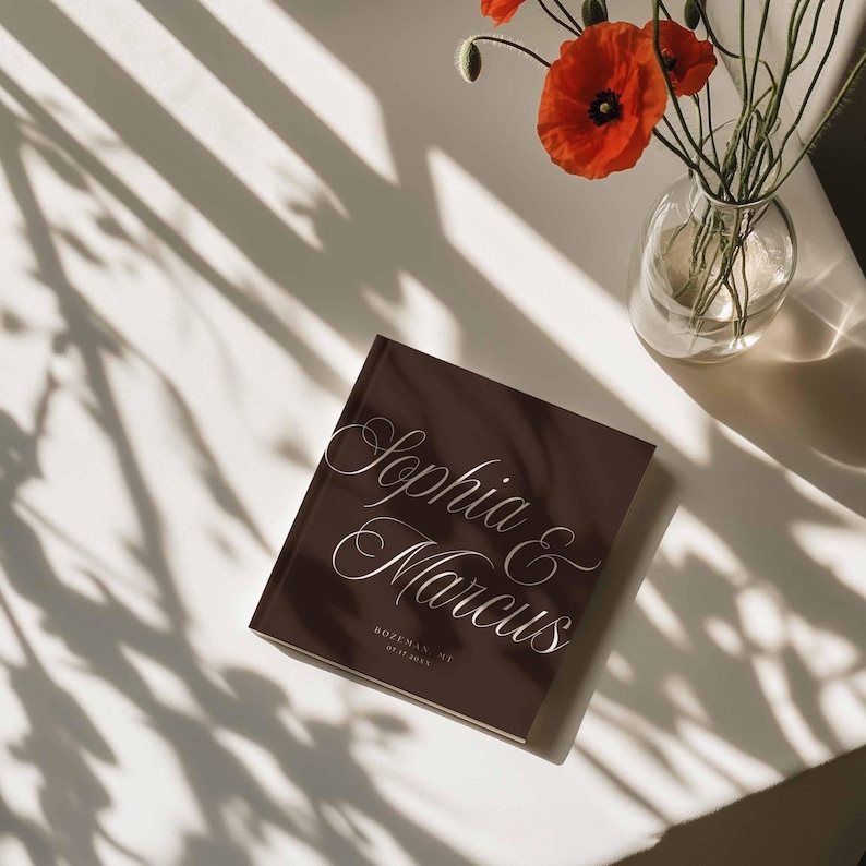 May include: A square dark brown book with white cursive text reading Sophia & Marcus, followed by the location Bozeman, MT and the date 07.15.20XX. The cover features a clean, minimalist design with elegant typography centered on the front surface.
