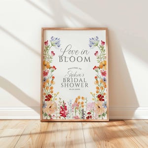 May include: A framed sign with the text "Love in Bloom" and "Welcome to Sophia's Bridal Shower" surrounded by a watercolor floral border. The flowers are in shades of red, orange, yellow, pink, and blue.
