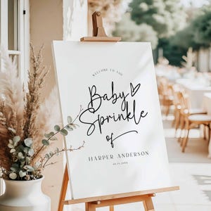 May include: A white sign on a wooden easel with the text "Welcome to the Baby Sprinkle of Harper Anderson" in black script. The sign is decorated with a small heart and floral accents. A vase of dried plants is in the foreground.
