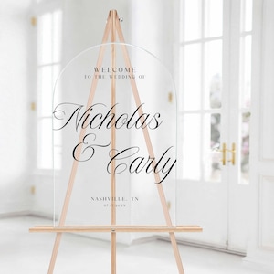 Pu&ograve; includere: Cartello di benvenuto per matrimoni in acrilico trasparente con una parte superiore ad arco, esposto su un cavalletto di legno. Il cartello presenta un'elegante scritta nera che recita "Benvenuti al matrimonio di Nicholas & Carly".