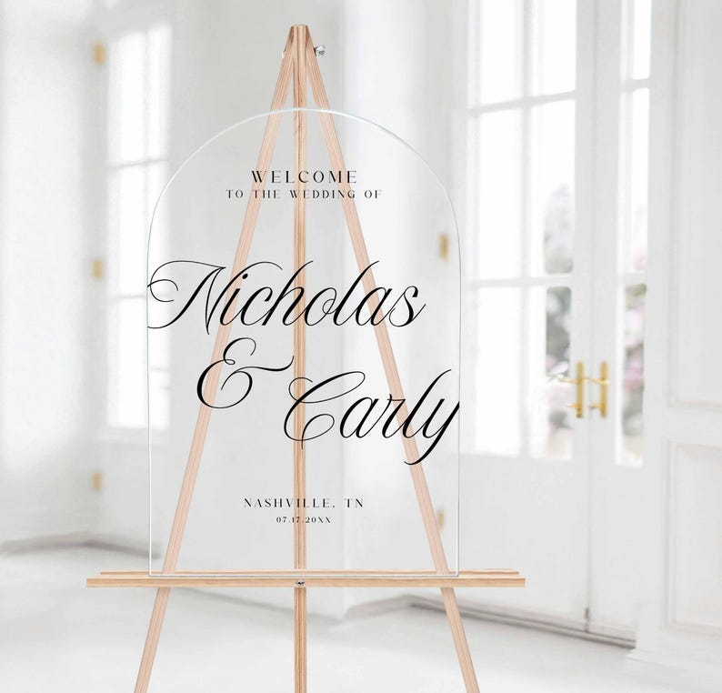 Pu&ograve; includere: Cartello di benvenuto per matrimoni in acrilico trasparente con una parte superiore ad arco, esposto su un cavalletto di legno. Il cartello presenta un'elegante scritta nera che recita "Benvenuti al matrimonio di Nicholas & Carly".