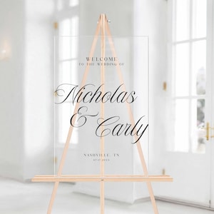 Pu&ograve; includere: Un cartello di benvenuto per matrimoni in acrilico trasparente su un cavalletto di legno. Il cartello presenta un'elegante scritta nera che recita "Benvenuti al matrimonio di Nicholas & Carly" con la localit&agrave; e la data sottostanti. Il cavalletto &egrave; in legno chiaro.