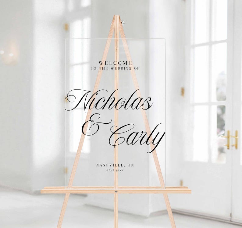 Pu&ograve; includere: Un cartello di benvenuto per matrimoni in acrilico trasparente su un cavalletto di legno. Il cartello presenta un'elegante scritta nera che recita "Benvenuti al matrimonio di Nicholas & Carly" con la localit&agrave; e la data sottostanti. Il cavalletto &egrave; in legno chiaro.