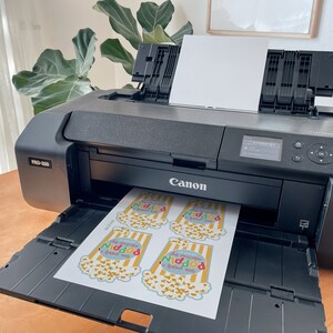 May include: A black Canon imagePROGRAF PRO-1000 printer with a sheet of paper in the output tray. The paper has four colourful popcorn bag designs printed on it, each with the text "This popcorn is good for your soul".