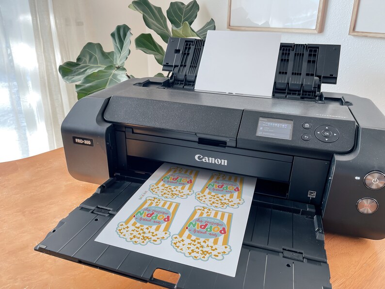 May include: A black Canon imagePROGRAF PRO-1000 printer with a sheet of paper in the output tray. The paper has four colourful popcorn bag designs printed on it, each with the text "This popcorn is good for your soul".