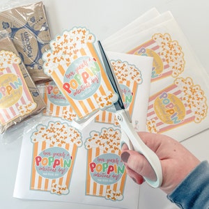 May include: A sheet of paper with several colourful popcorn-themed Valentine's Day stickers. The stickers feature a cartoon bucket of popcorn with the text "Have yourself a poppin' Valentine's Day!"  A hand is holding a pair of scissors and cutting out the stickers.