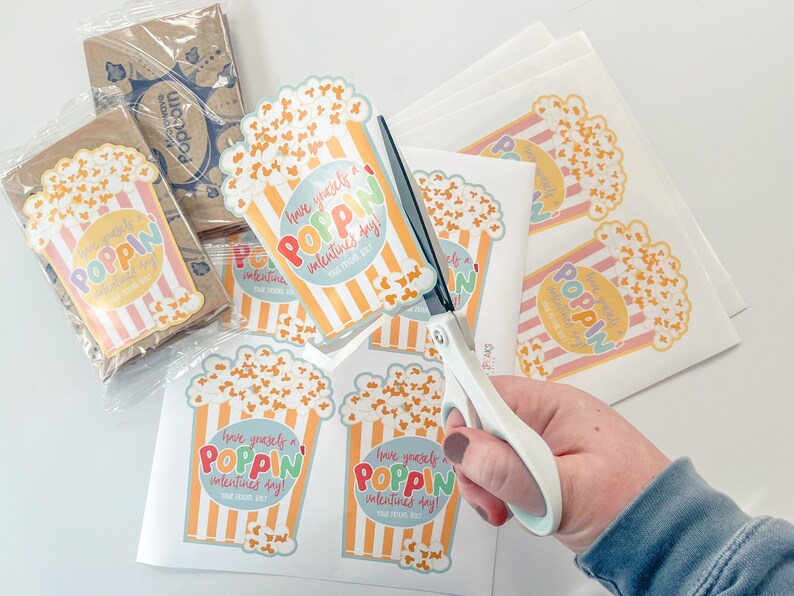 May include: A sheet of paper with several colourful popcorn-themed Valentine's Day stickers. The stickers feature a cartoon bucket of popcorn with the text "Have yourself a poppin' Valentine's Day!"  A hand is holding a pair of scissors and cutting out the stickers.