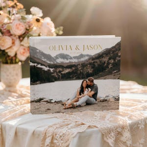 Peut inclure: Un album photo blanc avec l'inscription dor&eacute;e "OLIVIA & JASON" et "07.17.2023". La couverture pr&eacute;sente une photo panoramique d'un couple au bord d'un lac avec des montagnes en arri&egrave;re-plan. L'album est pos&eacute; sur une table avec une nappe en dentelle.