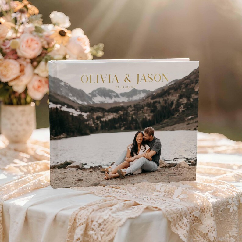 Peut inclure: Un album photo blanc avec l'inscription dor&eacute;e "OLIVIA & JASON" et "07.17.2023". La couverture pr&eacute;sente une photo panoramique d'un couple au bord d'un lac avec des montagnes en arri&egrave;re-plan. L'album est pos&eacute; sur une table avec une nappe en dentelle.
