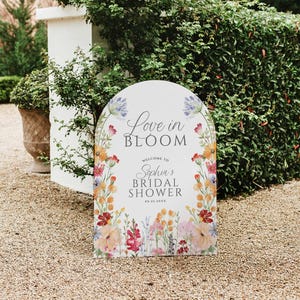 May include: A white, arched sign with floral accents and the text "Love in Bloom" and "Welcome to Sophia's Bridal Shower." The sign is decorated with colorful flowers in shades of pink, orange, yellow, and blue, perfect for a wedding or bridal shower.