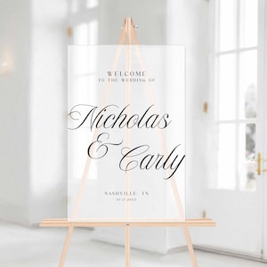 Pu&ograve; includere: Cartello di benvenuto per matrimoni in acrilico trasparente su un cavalletto di legno. Il cartello recita "Benvenuti al matrimonio di Nicholas & Carly" con la localit&agrave; e la data sottostanti. Il cartello &egrave; esposto in una stanza luminosa e ariosa.