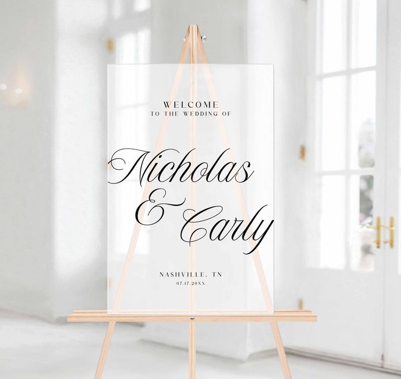 Pu&ograve; includere: Cartello di benvenuto per matrimoni in acrilico trasparente su un cavalletto di legno. Il cartello recita "Benvenuti al matrimonio di Nicholas & Carly" con la localit&agrave; e la data sottostanti. Il cartello &egrave; esposto in una stanza luminosa e ariosa.