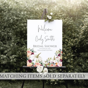 Op de afbeelding: Een wit bord op een zwart schildersezel, met de tekst "Welkom bij Carly Smith's Bridal Shower". Het bord heeft bloemen- en pompoenontwerpen. De datum is 26 september 2021. Het bord staat tegen een achtergrond van groen gebladerte.