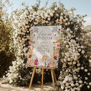 May include: A white sign on a wooden easel with the text "Love in Bloom" and "Sophia's Bridal Shower" surrounded by colorful floral illustrations. The sign is set against a backdrop of white roses and greenery.
