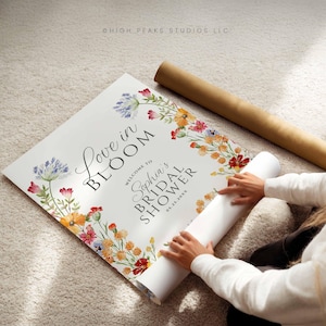 May include: A white bridal shower sign with floral accents and the text "Love in Bloom" and "Welcome to Sophia's Bridal Shower 05.25.20xx". The sign is partially rolled up, with a brown tube next to it.