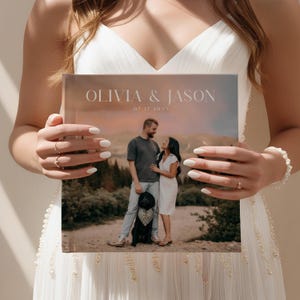 Peut inclure: Un album photo de mariage avec une photo d'un couple et de leur chien en ext&eacute;rieur. La couverture affiche "OLIVIA & JASON" et la date "07.17.20XX" en blanc. L'album est tenu par une personne portant une robe blanche, mettant en valeur le design et le contenu.