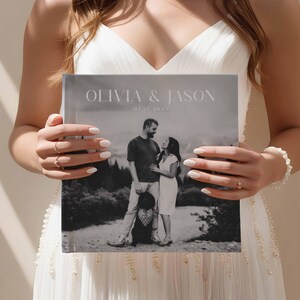 Peut inclure: Un album photo carr&eacute; avec les noms "OLIVIA & JASON" et la date "07.17.20XX" imprim&eacute;s sur la couverture. L'album pr&eacute;sente une photographie en noir et blanc d'un couple et d'un enfant dans un cadre naturel.