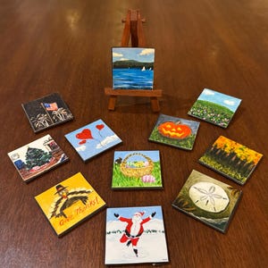 May include: A set of ten small, square paintings on wooden canvases. The paintings depict various scenes, including a sailboat on a lake, a pumpkin, a basket of Easter eggs, a turkey, Santa Claus, a seashell, a lighthouse, a heart-shaped balloon, a field of flowers, and a forest in autumn.