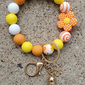 May include: A beaded keychain bracelet featuring a variety of colorful beads in yellow, orange, white, and patterned designs. The bracelet includes a gold-tone clasp, a flower charm, and a yellow tassel.