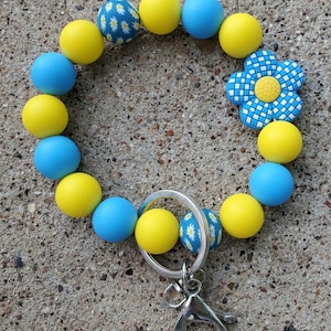 May include: A beaded keychain wristlet featuring yellow, blue, and daisy-patterned beads. The wristlet includes a silver-colored clasp and key ring. A blue and white flower-shaped charm is also included.