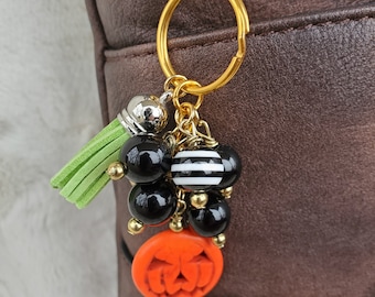 May include: A Halloween-themed keychain with a gold-colored clasp and key ring. The charm features black and white striped and solid black beads, a small green tassel, and an orange pumpkin face. The keychain is attached to a brown bag.