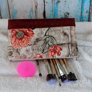 May include: A burgundy and beige floral makeup bag with a zipper closure. The bag is filled with a set of makeup brushes, including a pink makeup sponge.