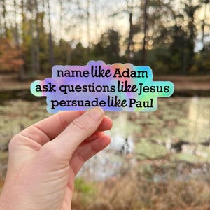 May include: A holographic sticker with the text "name like Adam ask questions like Jesus persuade like Paul". The sticker has a colorful, iridescent finish and is held up against a blurred outdoor background.