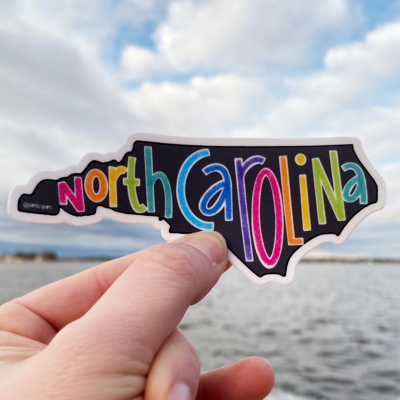 Może przedstawiać: Kolorowa naklejka z konturem stanu Karolina P&oacute;łnocna z napisem "north carolina" w kolorach tęczy. Naklejka jest trzymana w dłoni na tle niebieskiego nieba i wody.