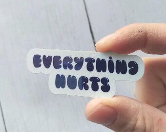 May include: A white sticker with the words "EVERYTHING HURTS" in dark blue bubble letters. The sticker is held between fingers against a light wood grain background.