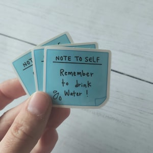 Puede incluir: Una nota adhesiva azul con el texto "Note to Self" y "Remember to drink water!" escrito en marcador negro.