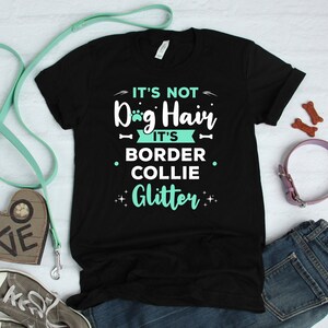 May include: Black t-shirt with the text "It's not Dog Hair, It's Border Collie Glitter" in turquoise lettering. The shirt is laid out on a white surface with a blue leash, a heart-shaped dog treat pouch, and a pink dog collar.