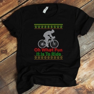 以下が含まれることがあります： 自転車に乗っている人のシルエットが描かれた、緑と赤のクリスマスをテーマにしたデザインの黒いTシャツ。Tシャツには「Oh What Fun It Is To Ride」という文字がプリントされています。