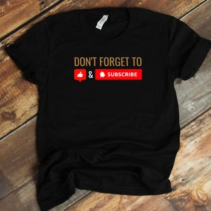 以下が含まれることがあります： 「DON'T FORGET TO」という金色の文字が書かれた黒いTシャツ。その下に赤い親指を立てた絵文字、アンパサンド、赤いベルの絵文字が並んでいます。「SUBSCRIBE」という文字は赤い背景に白い文字で書かれています。