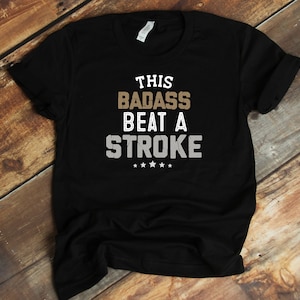 May include: Black t-shirt with the text "This badass beat a stroke" in white and brown lettering. The text has a distressed look. There are five stars below the text.