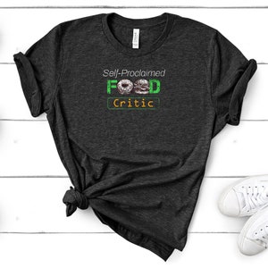 May include: A dark gray t-shirt with the text "Self-Proclaimed FOOD Critic" in green and orange. The word "FOOD" is illustrated with a donut and a hamburger.