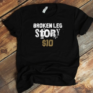 May include: Black t-shirt with white text that reads "BROKEN LEG STORY $10".