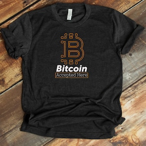 May include: A dark grey t-shirt with a white graphic of a Bitcoin symbol and the text "Bitcoin Accepted Here".
