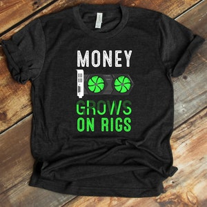 Puede incluir: Una camiseta gris oscura con el texto "MONEY GROWS ON RIGS" en blanco. Un gráfico de una tarjeta gráfica de computadora con dos ventiladores verdes está en el centro del texto.