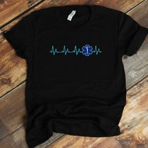 Op de afbeelding: Zwart T-shirt met een blauw en wit medisch symbool en een blauwe hartslaggrafiek. Het symbool is een ster van het leven met een slang eromheen.