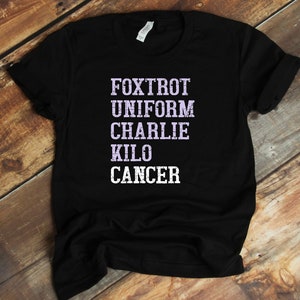 May include: Black t-shirt with the words "FOXTROT UNIFORM CHARLIE KILO CANCER" printed in white with a purple outline.