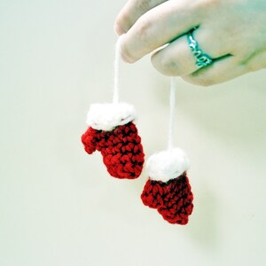 May include: Two small red and white crocheted mittens with white trim hanging from white strings. The mittens are held by a hand wearing a silver ring.