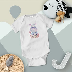 Puede incluir: Un body blanco para bebé con una ilustración acuarela azul y rosa de un hipopótamo sentado en un tronco con el texto 'cuddle bug'.