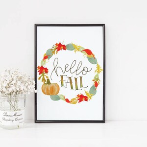 May include: A framed print with a white background and a wreath of fall leaves and pumpkins. The wreath surrounds the text "hello fall" in black script.