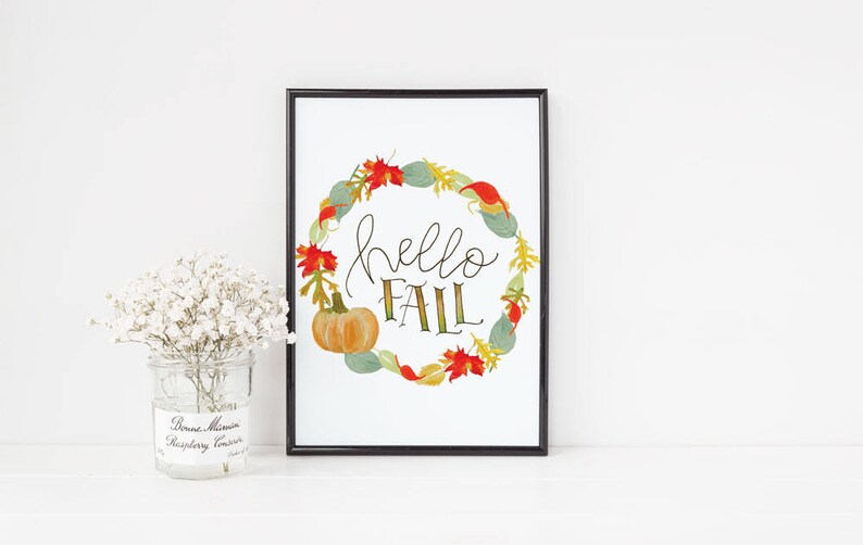 May include: A framed print with a white background and a wreath of fall leaves and pumpkins. The wreath surrounds the text "hello fall" in black script.